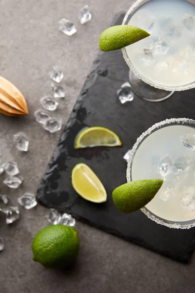 Top view of alcohol margarita cocktails with pieces of lime, ice cubes and wooden squeezer on grey tabletop — Stock Photo
