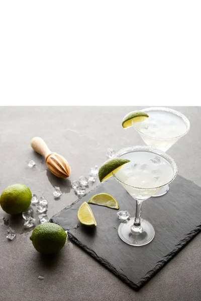 Vue rapprochée des cocktails margarita avec des morceaux de lime et un pressoir en bois sur la surface grise sur blanc — Photo de stock