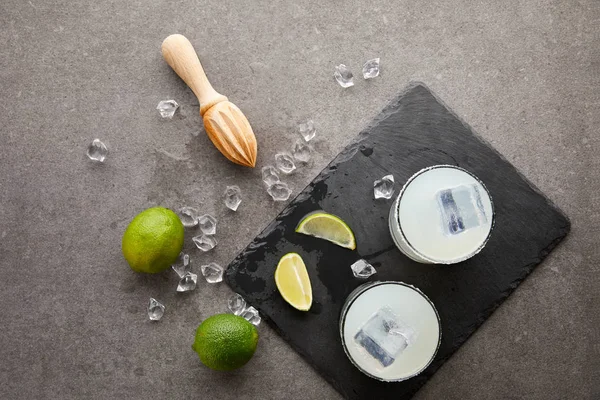 Flat lay with wooden squeezer, refreshing sour caipirinha cocktails with lime and ice on grey tabletop — Stock Photo