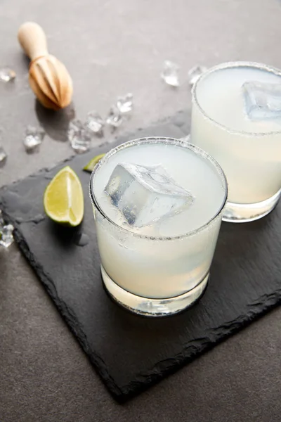 Close up view of wooden squeezer, refreshing sour caipirinha cocktails with lime and ice on grey tabletop — Stock Photo