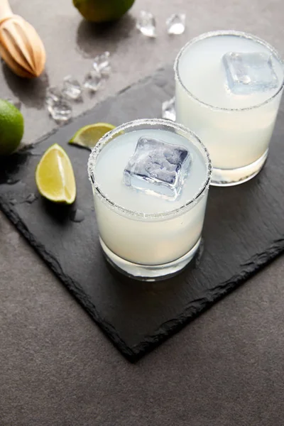 Close up view of wooden squeezer, refreshing sour caipirinha cocktails with lime and ice on grey tabletop — Stock Photo