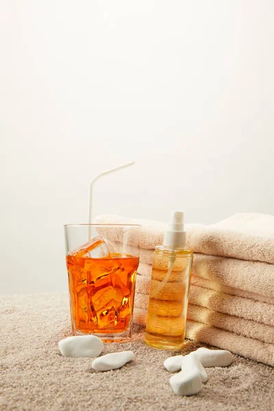 Vue rapprochée de l'huile de bronzage, cocktail rafraîchissant avec glace et paille et serviettes sur sable sur fond gris — Photo de stock