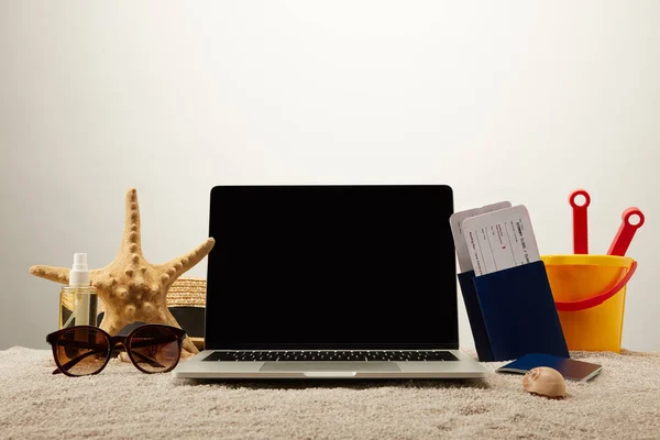 Nahaufnahme von Laptop mit leerem Bildschirm, Seestern, Sonnenbrille, Reisepässen mit Eintrittskarten und Spielzeugeimer auf Sand auf grauem Hintergrund — Stockfoto