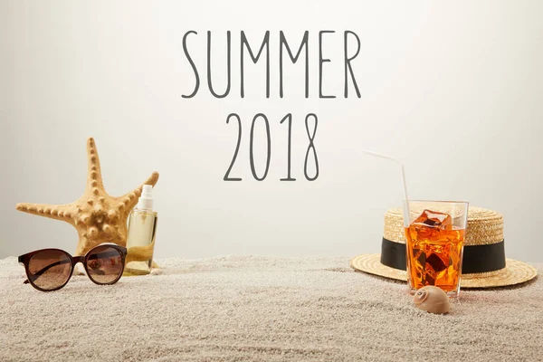 Close up view of summer 2018 lettering, cocktail with ice, straw hat, sunglasses and tanning oil on sand on grey backdrop — Stock Photo