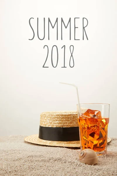 Close up view of summer 2018 lettering, straw hat and refreshing cocktail on sand on grey backdrop — Stock Photo