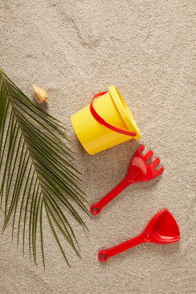 Tendido plano con juguetes de colores, concha marina y hoja de palma verde en la arena - foto de stock