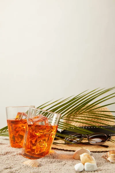 Крупный план соломенной шляпы, коктейлей, ракушек, солнцезащитных очков и пальмового листа на песке на сером фоне — стоковое фото