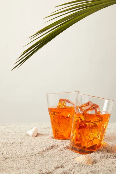 Close up view of cocktails with ice with palm leaf and seashells on sand on grey background — Stock Photo