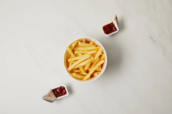 Vista superior de batatas fritas em tigela com recipientes de ketchup em branco — Fotografia de Stock