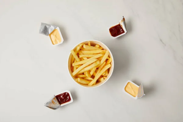 Top view of tasty french fries in bowl surrounded with containers of sauces on white — Stock Photo