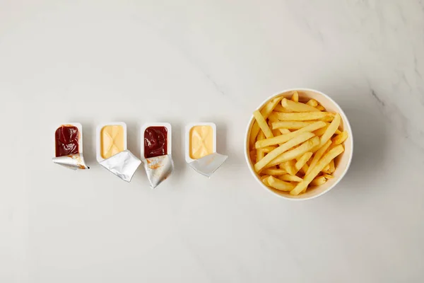 Vue du dessus des frites dans un bol avec des récipients de sauces en rangée sur blanc — Photo de stock