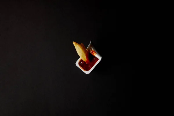 Vista dall'alto del contenitore di plastica con ketchup con patate fritte versate isolate sul nero — Foto stock