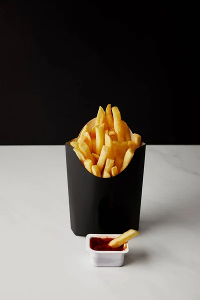 Scatola di patatine fritte con contenitore di plastica di ketchup su superficie di marmo bianco isolato su nero — Foto stock