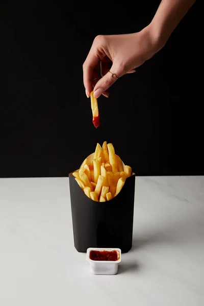 Colpo ritagliato di donna pieghevole friggere francese versato nel ketchup sopra scatola di patatine fritte isolate su nero — Foto stock