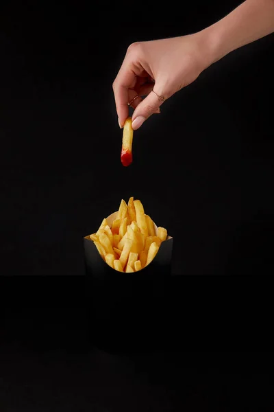 Colpo ritagliato di donna pieghevole friggere francese versato nel ketchup sopra scatola di patatine fritte isolate su nero — Foto stock