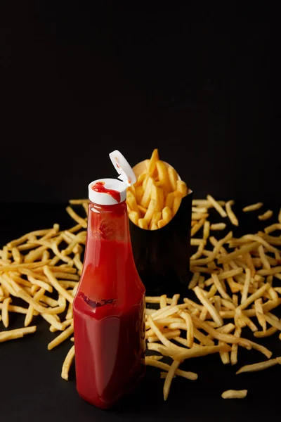 Bottle of ketchup with messy french fries on black surface isolated on black — Stock Photo