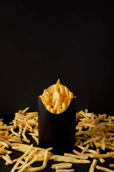 Close-up shot of delicious french fries in black box surrounded with messy fries on tabletop isolated on black — Stock Photo