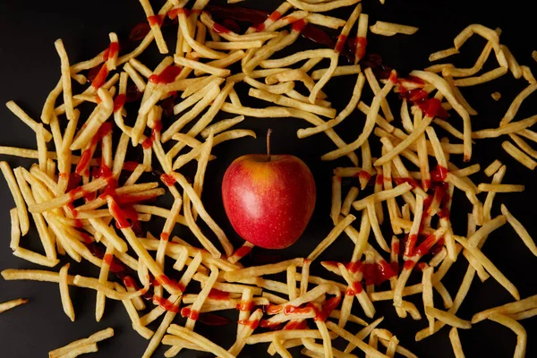 Top view of red apple surrounded with french fries poured with ketchup isolated on black — Stock Photo
