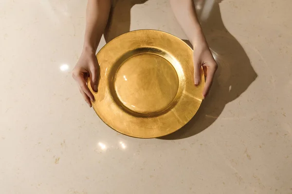 Золота металева тарілка в жіночих руках на світлому столі — стокове фото
