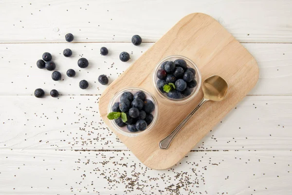 Vue de dessus des puddings de chia avec des bleuets frais et de la menthe sur planche à découper en bois sur surface blanche — Photo de stock