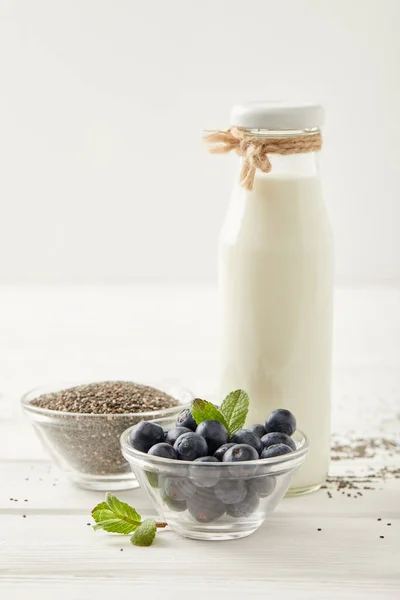 Vista da vicino di bottiglia di latte, mirtilli e semi di chia per fare il budino di chia su un tavolo di legno bianco — Foto stock