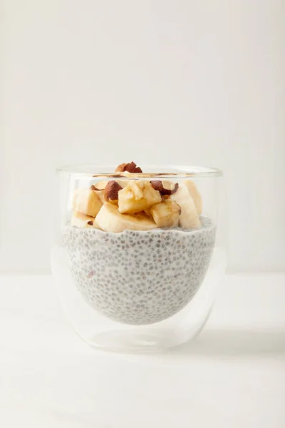 Vue rapprochée du savoureux pudding aux graines de chia avec des morceaux de banane et de noisettes sur le dessus de table blanc — Photo de stock