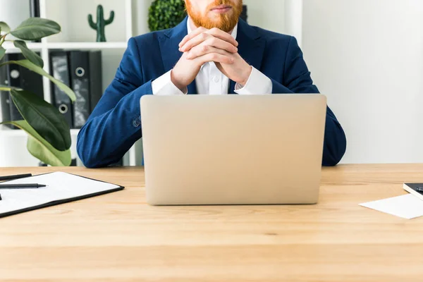 Vista parcial del hombre de negocios barbudo en traje sentado en el lugar de trabajo con el ordenador portátil en la oficina - foto de stock
