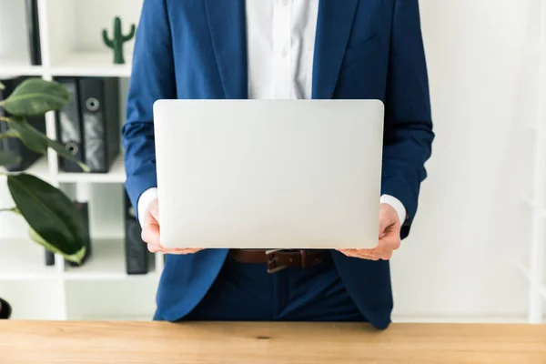 Tiro cortado de homem de negócios no terno que mantém o computador portátil em mãos no local de trabalho no escritório — Fotografia de Stock