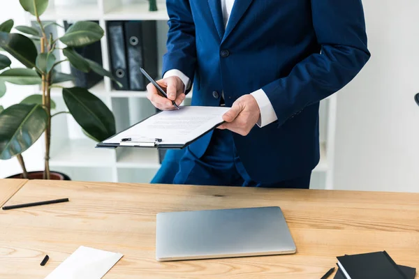 Обрезанный снимок бизнесмена с блокнотом и ручкой в руках на рабочем месте с ноутбуком в офисе — стоковое фото