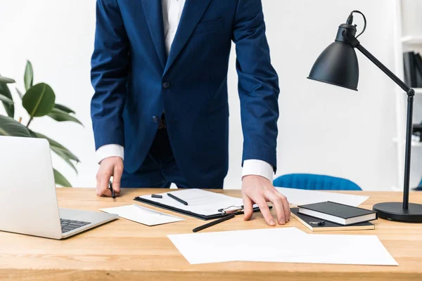 Vue partielle de l'homme d'affaires en costume sur le lieu de travail avec des cahiers, des documents et un ordinateur portable au bureau — Photo de stock