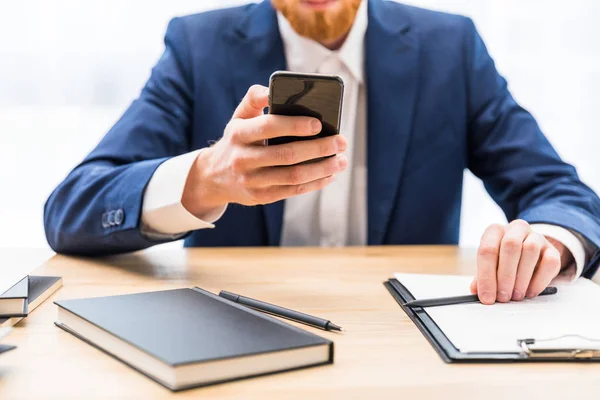 Vista parcial del hombre de negocios en traje usando teléfono inteligente en el lugar de trabajo con portátil en la oficina - foto de stock