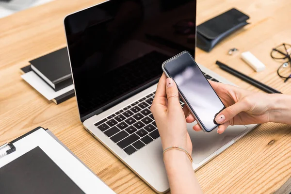 Обрезанный снимок бизнесвумен с помощью смартфона на рабочем месте с ноутбуком — стоковое фото