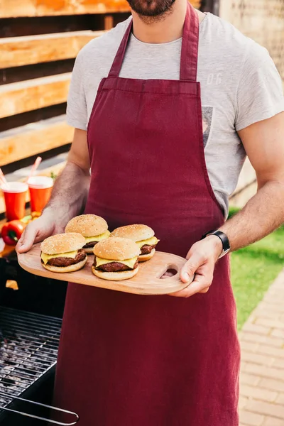 Close-up view of man holding board with burgers cooked outdoors on grill — Stock Photo