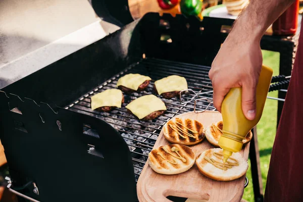 Chef adding mustard to burgers cooked outdoors on grill — Stock Photo