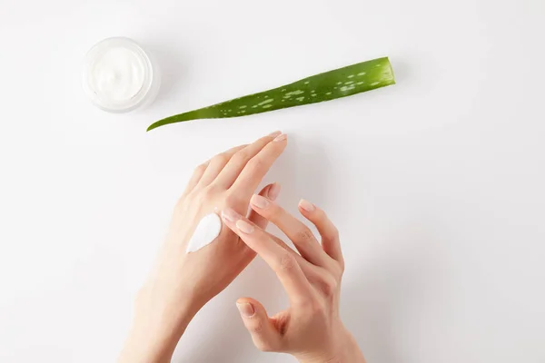 Plan recadré de femme appliquant de la crème biologique sur les mains, feuille d'aloe vera et récipient à crème sur la surface blanche — Photo de stock