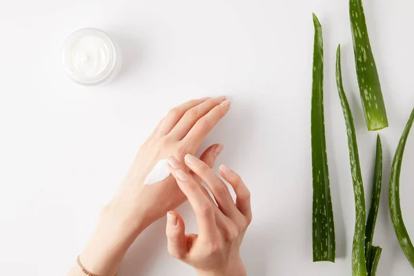 Cropped image of woman applying cream on hands, aloe vera leaves and cream in container on white surface — Stock Photo