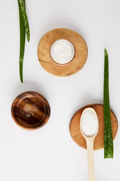 Vista elevada de creme orgânico em recipiente e colher com sal em fatias de madeira, folhas de aloe vera e tigela de madeira com suco de aloe vera na superfície branca — Fotografia de Stock