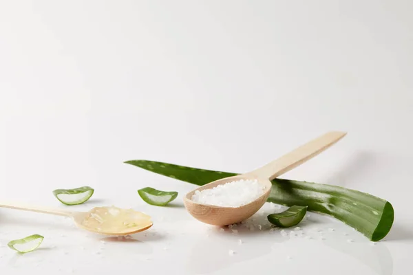 Vue rapprochée de deux cuillères en bois avec du jus et du sel d'aloe vera, une feuille d'aloe vera et des tranches sur une surface blanche — Photo de stock