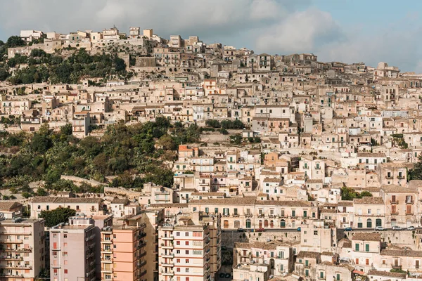 Luz del sol en casas cerca de árboles verdes contra el cielo con nubes en ragusa, italia - foto de stock