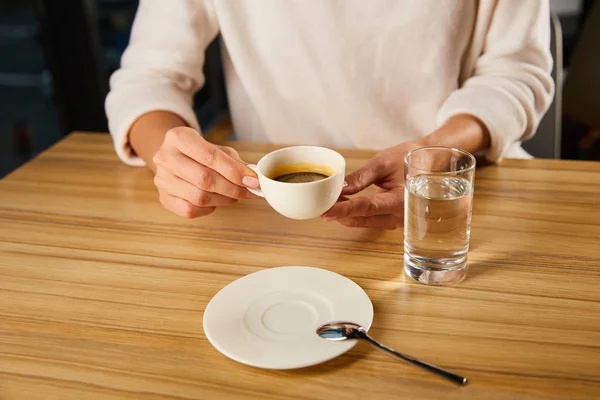 Cropped view of woman holding cup of hot coffee near glass of fresh  water in cafe — Stock Photo