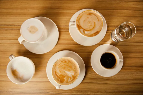 Top view of saucers with cups of different coffee near glass of water — Stock Photo