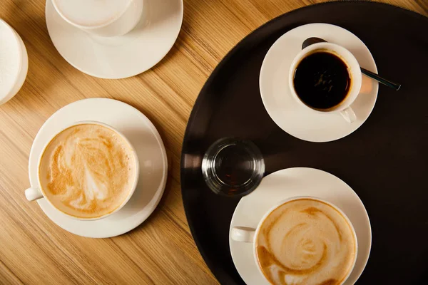 Vista dall'alto di tazze di caffè diverso vicino a vetro d'acqua su vassoio — Foto stock