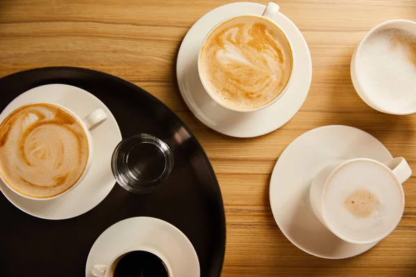 Vista superior de tazas de café diferentes cerca de vaso de agua en bandeja negra - foto de stock
