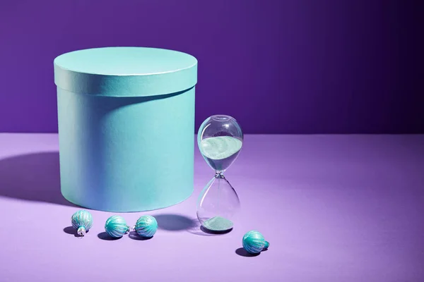 Boîte cadeau bleu et boules décoratives et sablier sur fond violet — Photo de stock