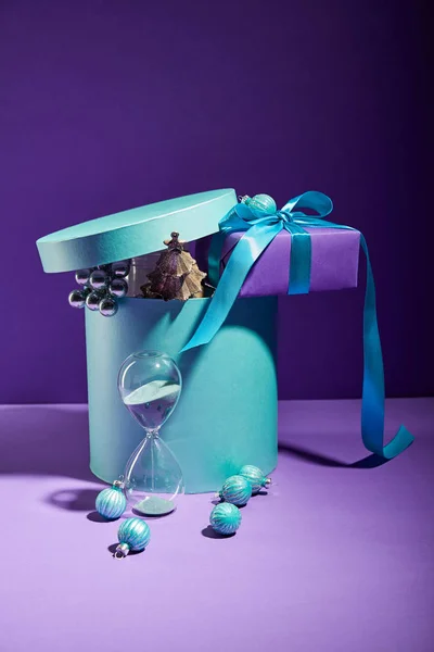 Blue box Christmas decoration and present near hourglass on purple background — Stock Photo