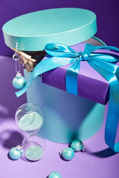 Selective focus of blue box Christmas decoration and present near hourglass on purple background — Stock Photo