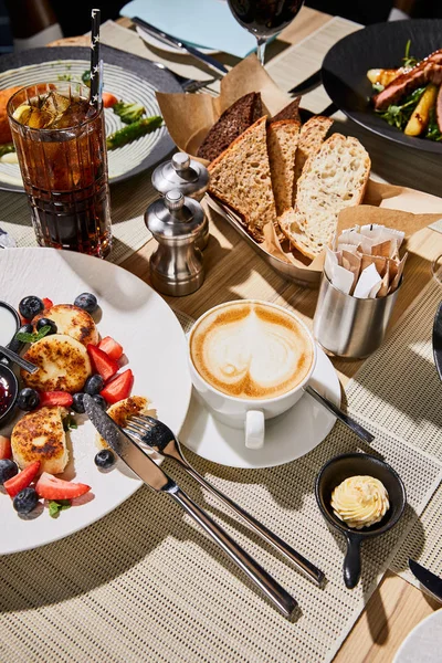 Mesa com pratos de restaurante e panquecas de queijo com bagas perto de cappuccino — Fotografia de Stock