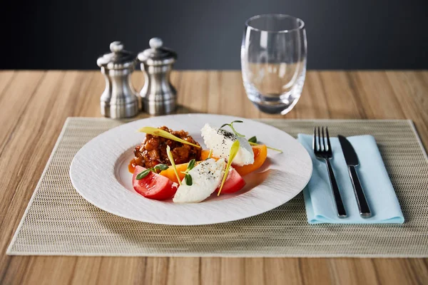 Foyer sélectif de délicieux plat de restaurant avec caviar d'aubergine et tomates servi sur une table en bois avec de l'eau, salière et poivrière et couverts isolés sur gris — Photo de stock