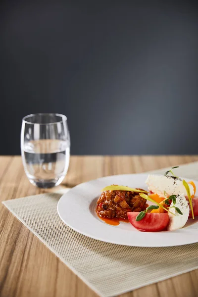 Селективный фокус изысканного ресторанного блюда с баклажанной икрой и помидорами, подаваемых на деревянном столе с водой, изолированной на сером — стоковое фото