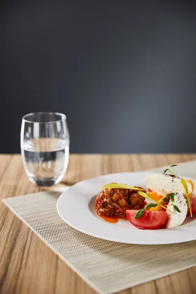 Foco seletivo de delicioso prato de restaurante com caviar de berinjela e tomates servidos em mesa de madeira com água isolada em cinza — Fotografia de Stock